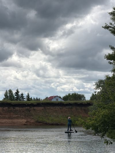 The image depicts a solitary individual paddleboarding on a placid river in Il'inka, Russia, under an overcast sky. The sky, dominating the upper two-thirds of the frame, is filled with dense, uniform grey and white clouds. The river occupies the lower quarter, its surface subtly rippled, reflecting the muted light.
In the mid-ground, a person stands on a dark-colored paddleboard, facing away from the viewer and towards the distant riverbank. The individual is dressed in a dark teal or green jacket, dark pants, and a light-colored head covering, actively propelling the board with a black paddle held on the right side.
Behind the paddleboarder, a steep, eroded riverbank rises from the water. Its lower section reveals layers of reddish-brown earth and sand, while the upper portion is covered with green grass and varied vegetation. Along the top of the bank, a line of trees extends across the scene. On the left, several coniferous trees are visible, while on the right, deciduous trees and bushes are present. Partially obscured by trees on the upper right, a single-story house with a reddish-brown gabled roof and light-colored siding is situated on the elevated land. A portion of a tree with green foliage extends into the foreground on the right, partially framing the scene.