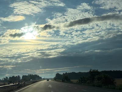 The image depicts a highway scene in Zagore, Bulgaria, under a dramatic sky filled with a mix of bright sunlight breaking through layered clouds and darker, heavier cloud formations. The sun is a prominent feature, casting a bright glare and visible rays towards the left side of the frame. The highway stretches into the distance, appearing empty except for a single black car visible further down the road, heading away from the viewer. The road is bordered by a metal guardrail on the left, with reflective markers. A line of dense trees forms a dark silhouette along the horizon, separating the road from the sky. The overall impression is one of a journey through a somewhat overcast but brightening landscape.