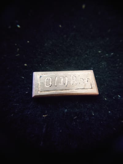 A rectangular metal tag with the inscription "0(I)Rht" is placed on a dark, textured surface. The tag is made of a light-colored metal, possibly copper or brass, and is slightly beveled on its edges. The inscription appears to be etched or stamped into the surface of the tag. The dark background is out of focus and appears to be fabric or a similar material, with small specks scattered across it. The lighting is dim, casting a slight shadow on the surface beneath the tag. The object is centrally located in the frame. There are no people or other objects visible in the image. There is no information that can place this object in Sokolohirsk, Ukraine.