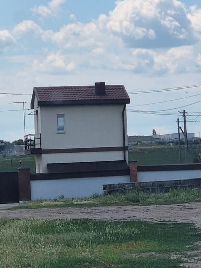 The image shows a two-story building with a brown tiled roof and light beige walls. A small balcony with dark metal railings is attached to the second story. A single rectangular window is visible on the second story. The building is bordered by a white wall with a brown trim, and a dark metal gate is partially visible on the left. A brick pillar stands next to the gate. The foreground features green grass and a dirt path. In the background, there are power lines, a utility pole, and some industrial buildings. The sky is blue with scattered white clouds. The scene appears to be in an urban or suburban area, possibly on the outskirts of Pidhaitsi, Ukraine, given the context. There are no people visible in the image.