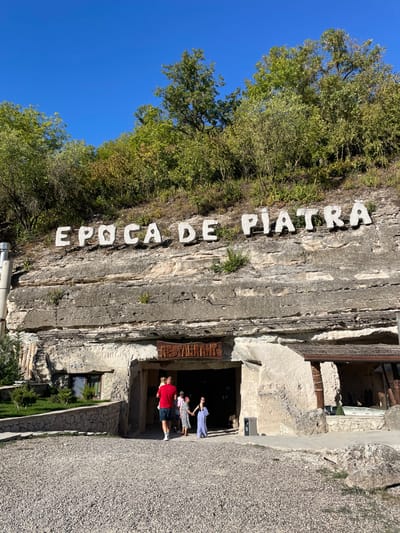This image depicts the "Epoca de Piatra" (Stone Age) complex, located in the city of Brănești, Moldova. The scene is outdoors on a clear, sunny day.

The primary subject is a building carved into a rock face or cliff, featuring a large, central entrance. Above this structure, large white letters spelling "EPOCA DE PIATRA" are affixed to the cliff face. Directly above the main entrance, a rustic wooden sign displays the word "RESTAURANT."

A group of approximately five individuals, including adults and children, are positioned at or near the entrance. A male adult, wearing a red t-shirt and dark shorts, is observed carrying a small child in his arms, facing away from the camera and towards the entrance. Two other children, one in a light-colored dress and another in a patterned dress, are walking hand-in-hand towards the entrance. Another adult female is partially visible behind them.

The ground in the foreground is covered with light-colored gravel, forming a path leading to the entrance. To the left of the entrance, a small landscaped area with green bushes and a single window is visible. A metal chimney or ventilation pipe runs vertically along the left side of the rock face. To the right of the entrance, an open-sided structure with a wooden roof and beams is present, suggesting an outdoor seating area or extension. The top of the cliff is densely covered with green trees and foliage under a bright blue sky.