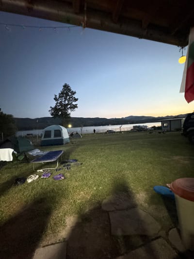 At dusk, a campsite is set up near the lake in Golyama Zhelyazna, Bulgaria. The sky is a gradient of deep blue and pale orange, with a single tree silhouetted against the horizon. Several tents are pitched on the grassy ground, along with camping chairs and a folding table. Various pairs of footwear, including flip-flops and clogs, are scattered on the grass. In the distance, a few vehicles and structures are visible near the water's edge. A Bulgarian flag hangs from an overhang in the foreground. The scene suggests a relaxed evening gathering at a campground by a lake.