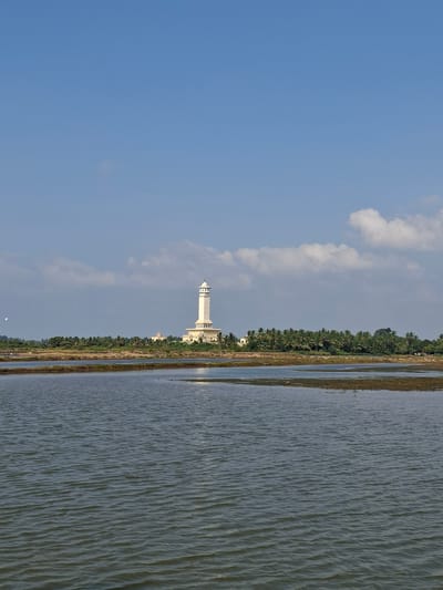 The image depicts a landscape with a lighthouse and a body of water. The sky is a clear blue with scattered white clouds, suggesting it's daytime with fair weather. A tall, white lighthouse with a distinctive top is visible in the mid-ground, situated behind a line of lush green trees. These trees form a dense border between the lighthouse and a calm, rippling body of water that occupies the right side of the frame. The water's surface has a textured appearance due to small waves, reflecting the sky and the surrounding landscape. A thin strip of dry land or vegetation is visible between the trees and the water. The overall scene conveys a sense of tranquility and natural beauty, possibly a coastal or lakeside setting. The location is identified as Dayahtuha, Indonesia. There is no text visible in the image.