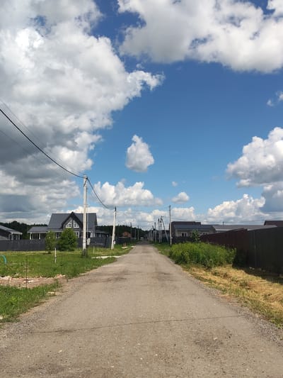 This is an eye-level, medium shot of a gravel road in the village of Skurygino, Russia, on a sunny day. The road leads into the distance, lined on both sides by houses and fences. On the left, a dark-colored house with a gray roof and white trim is visible behind a dark gray fence. Further down the road on the left, more houses with fences can be seen. On the right side of the road, there are green bushes and tall grass, followed by a dark fence. The sky is a bright blue, dotted with fluffy white clouds. Power lines with poles are present along the road. The environment appears to be a residential area in a rural setting.