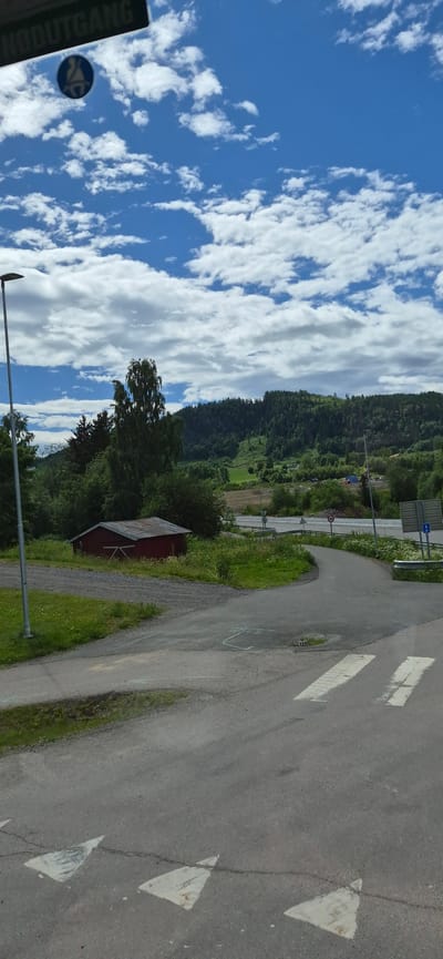 This media file shows a scenic view with a road and a blue sky filled with white clouds. The road is paved and curves to the right, with white triangular markings and some faint white arrow markings painted on it. To the left of the road, there is a grassy area with a small red building. Beyond the building and the road, a lush green forest covers a hillside. In the distance, there are signs and what appears to be a traffic light. The sky is a vibrant blue with fluffy white clouds, suggesting a clear day. The overall setting is rural and serene, possibly in a mountainous or hilly region, consistent with the location context of Biri, Norway. The lighting indicates it is daytime. There is no visible activity or people in the frame. Some text is visible on a sign on the far left, which reads "UTGANG".
