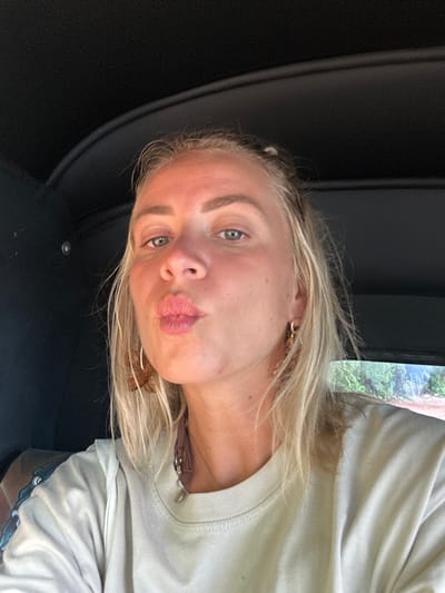 A blonde woman with light skin is facing the camera with pursed lips. She has blue eyes and wears delicate gold earrings. She is in what appears to be a car, judging by the dark interior and the glimpse of green foliage outside the window. The woman is wearing a light-colored t-shirt. A necklace is visible around her neck, partially obscured by the shirt.