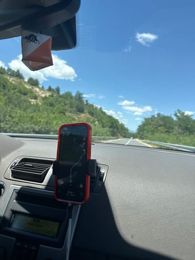 In the interior of a car, a red smartphone is mounted on the dashboard, displaying a navigation application with a route highlighted. The phone is positioned in front of a car's air vent and above the car's audio system, which shows "AUX" and "Audio Input" on its display. A small fabric flag, with a black silhouette of a creature and orange and white geometric patterns, hangs from the rearview mirror. The exterior view through the windshield shows a road winding through a lush, green, hilly landscape under a bright blue sky with scattered clouds. The environment suggests a scenic drive on a sunny day in a rural or natural setting, possibly on a road leading to or from the city of Sadovo, Bulgaria, given the context.
