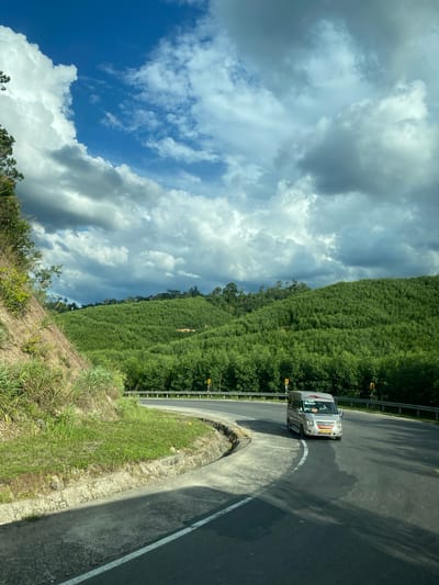 A silver van with "LUXURY" written on its side drives on a paved road that curves to the right, bordered by a concrete curb and a metal guardrail. The road is flanked by a steep, grassy embankment on the left and a dense, green forest covering rolling hills on the right. The sky above is a vibrant blue with dramatic, dark gray clouds casting shadows. The image appears to be taken from a moving vehicle, looking towards the van and the scenic landscape. The location is identified as Thôn 18, Vietnam.