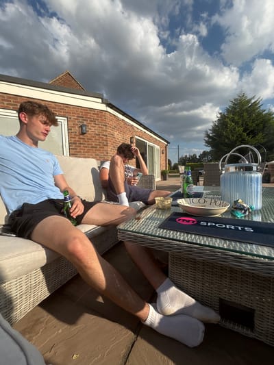 Two young men relax on a patio. One in a blue shirt and black shorts lounges with a beer. The other sits behind him, adjusting his hair. A glass-topped wicker table holds drinks, a bucket, and promotional items. White socks are seen under the table. A brick house stands in the background beneath a cloudy sky.