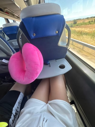 The image shows the interior of a bus with seats and a window. A bright pink neck pillow is placed on a gray tray table attached to the back of a blue and gray bus seat. The pillow is positioned in the foreground. To the right of the pillow, a person's legs are visible; they are wearing white shorts. To the left of the pillow, another person's legs are visible, wearing black shorts with a white stripe and writing. The window on the right side of the bus shows a blurred landscape of a field and trees under a bright sky, indicating the bus is in motion. The seats appear to be for public transportation, possibly a tour bus or intercity bus. The scene is observed from a perspective slightly above and to the side of the neck pillow and the seated passengers. The location in Shtarkovo, Bulgaria is not discernible from the visual information provided in the image.