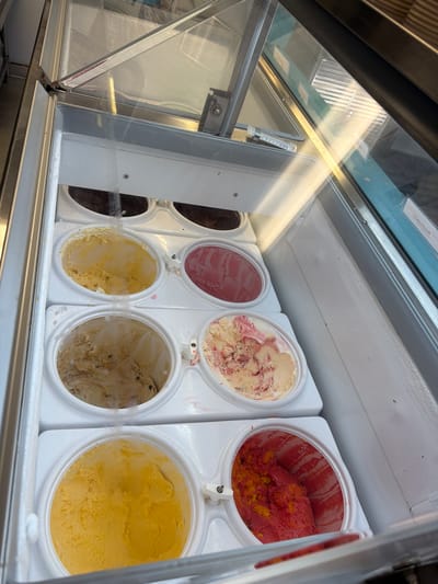 The image provides an overhead view into a commercial ice cream display freezer, likely situated in an ice cream shop or cafe in Healy, United States. The freezer contains multiple round white containers filled with various flavors of ice cream.

The scene showcases a selection of colorful frozen desserts. In the top row, from left to right, there is a dark brown (likely chocolate), a solid pink/red flavor, a creamy white ice cream with red swirls (possibly strawberry or raspberry ripple), and a vibrant red and yellow mixture, resembling a fruit sorbet or sherbet. The bottom row features a partially visible dark brown flavor, a pale yellow, a light brown ice cream with visible chunks (such as cookies, nuts, or chocolate pieces), and a bright, uniform yellow flavor (like lemon or mango).

Some glass lids of the freezer are open, while others are closed, reflecting ambient light from above. Small, rectangular white labels are attached to the glass covers, indicating the flavors. Visible text on these labels includes "STRAWBERRY CHEESECAKE" and possibly "Golden Chocolate". A clear plastic object, potentially a thermometer or a label holder, is mounted on the metal frame of the freezer. The overall presentation suggests a well-stocked and ready-to-serve ice cream selection. No people or specific activities are visible, just the static display of the frozen treats.