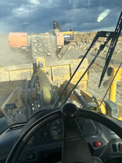 The image shows the interior of a heavy machinery vehicle, likely a wheel loader or similar earthmoving equipment. The view is from the driver's seat, looking through the windshield. The steering wheel and dashboard are prominent in the foreground. Through the windshield, there's a view of other construction equipment operating in a quarry or construction site. The environment is dusty, and there's evidence of recent earthmoving activity. The location is reported to be Hristiyanovo, Bulgaria.