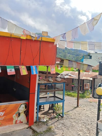 The image depicts a roadside establishment in Tharpu, Nepal, under a partly cloudy sky. A bright red building forms the backdrop, adorned with strings of colorful prayer flags and miniature national flags, including those of Mexico, Spain, and Argentina. A young woman is depicted in an advertisement on the building's lower section, holding an ice cream. Adjacent to the red building is a small blue metal structure, possibly a food stall or display unit, with shelves and some debris around its base. Further back, several corrugated tin-roofed shacks are visible, along with lush green hills in the distance. The ground is covered with gravel and scattered vegetation.