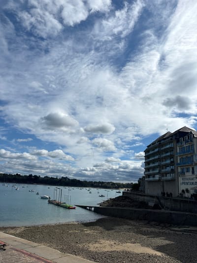 The image shows a view of the harbor in Dinard, France, taken from a waterfront promenade. The coordinates (latitude: 48.636, longitude: -2.049) correspond to a location near the "Le Printania" restaurant. The scene features a clear blue sky with white fluffy clouds and a calm body of water in the foreground. There are many small boats scattered across the water. A stone wall with a metal railing runs along the edge of the harbor, separating the promenade from the water. A multi-story building with balconies and blue accents can be seen in the background. The building has a sign for a restaurant called "Le Printania" on its side. There are a few people walking along the promenade on the right side of the image. The promenade is covered in small pebbles. The image was taken during a sunny day.