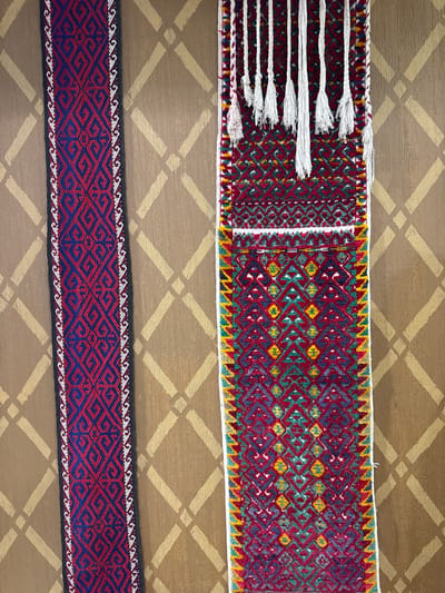 Two pieces of intricately woven textiles are displayed vertically against a wall with a textured, patterned background. The textile on the left is a narrow band with a repeating geometric pattern in red and dark blue, bordered by white and black trim. The textile on the right is wider and features a more complex pattern of red, green, yellow, and blue, with white tassels hanging from the top edge. The wall behind the textiles has a diamond-shaped stencil pattern in a lighter beige color against a darker brown background. This scene is located in Fort-Şevtşenko, Kazakhstan.