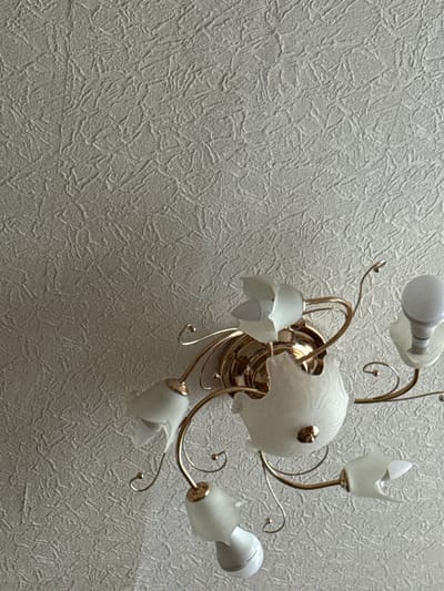 This media file shows a close-up of a ceiling light fixture mounted on a textured wallpapered ceiling. The light fixture is made of gold-toned metal and has several arms extending from a central decorative piece. Each arm holds a frosted white glass shade shaped like a flower or a tulip, with some having visible light bulbs inside. One of the bulbs is a white LED bulb, while another appears to be an incandescent bulb with a rounded top. The central part of the fixture has a larger, globe-like frosted glass element with a gold finial. The wallpaper has a distinctive rough, ridged texture in a neutral gray or off-white color, creating a visually interesting background. The lighting suggests an indoor setting, possibly a home or a room in a building. The image is a still shot with no apparent activity taking place; it is a static depiction of the ceiling fixture. There is no visible text in the image. The context of Piatykhatky, Ukraine, places this scene within a specific geographic location.