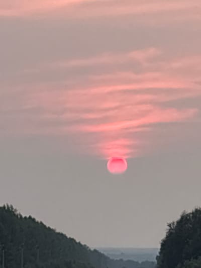 A wide, eye-level shot captures a hazy, pink sunset over Staryi Poros, Russia. The sun, a bright red orb, is partially obscured by soft, wispy clouds that streak horizontally across the pale pink sky. In the foreground, a dark, sloping hill covered in dense trees lines the bottom of the frame on the left, while a similar, though less defined, wooded area is visible on the right. The landscape suggests a rural or semi-rural setting, possibly near a road or highway given the subtle hints of vertical structures on the left hillside. The overall atmosphere is serene and slightly melancholic, dominated by the soft, diffused light of the setting sun.