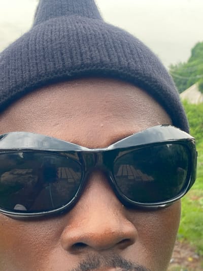 A close-up, rotated view of a person wearing a dark beanie and sunglasses. The person has dark skin and a beard, and their nose is prominently featured in the frame. The sunglasses reflect a scene outside, possibly a building and greenery. The background suggests an outdoor setting, though the exact location in Amaigbo, Nigeria is not discernible from this image.