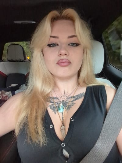 A blonde female is seated in a vehicle, depicted from the chest up. She has light blue eyes with dark winged eyeliner and pink lipstick on slightly pursed lips. Her long, wavy blonde hair, styled with bangs, frames her face. She wears a black, V-neck, sleeveless top featuring metal grommets and button-like closures down the front. A prominent chest tattoo of a stylized winged insect with a sunburst design is visible on her sternum. Two gold-chain necklaces are worn: one with a blue-green stone pendant shaped like a dragonfly or butterfly, and another with a clear to smoky quartz crystal point pendant. A gray seatbelt crosses diagonally over her right shoulder and chest. The vehicle's interior is dark, with a light-colored headliner visible overhead and a light-colored seat back to her right. A dark headrest is visible behind her on the left, and through the window behind it, green foliage and a partial yellow road sign are visible, indicating an outdoor, possibly rural, environment. The specific location within Electoral Area A, Canada, or any city therein, cannot be identified from the visual information provided.
