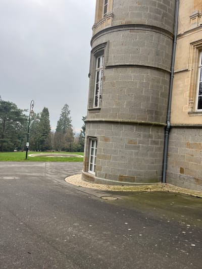 The image captures an outdoor scene in Bagnoles de l'Orne Normandie, France, under an overcast sky. The foreground consists of dark asphalt pavement, showing scattered light-colored specks and two circular metal manhole covers. To the right, a multi-story cylindrical tower, constructed from grey rectangular stone blocks, dominates the midground. It features several white-framed, multi-paned windows. A curved section of light-colored cobblestone pavers borders the base of the tower where it meets the asphalt. A grey vertical downspout runs along the right side of the tower, connecting it to an adjacent building section with lighter, yellowish-cream colored walls and more ornate stone detailing around its windows. To the left, a manicured green lawn extends into the background. A black streetlamp with an ornate, curved top and an attached white sign stands on the grass. Further back on the lawn, a circular, low-walled structure, possibly a dry fountain basin, is visible. The background is composed of a dense line of trees, including tall conifers and leafless deciduous trees, beneath a uniform light grey sky. No people or active subjects are present.