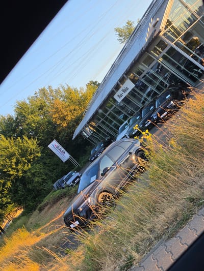 In Bischofsheim, Germany, at a car dealership, a row of cars is displayed on a grassy incline in front of a modern building with a glass facade. The cars, primarily black and silver, are parked neatly in a line. One prominent black station wagon is in the foreground, with its front wheel visible and a "Max" logo on its license plate frame. Beyond it, other vehicles, including a silver Audi, are also on display. A white flag with a red and blue logo and text reading "XAM" is planted in the grass to the left of the cars. The dealership building, identified by a "Max-Weiss" sign on its exterior, features large glass windows that reflect the surrounding environment and the sky. Tall green trees with some yellowing leaves flank the dealership and the parked cars, suggesting it might be autumn. The scene is bathed in the warm, golden light of a setting sun, casting long shadows and highlighting the dry grass. The ground in the foreground is paved with interlocking grey stones. The image captures a typical outdoor car lot setting in a German town.