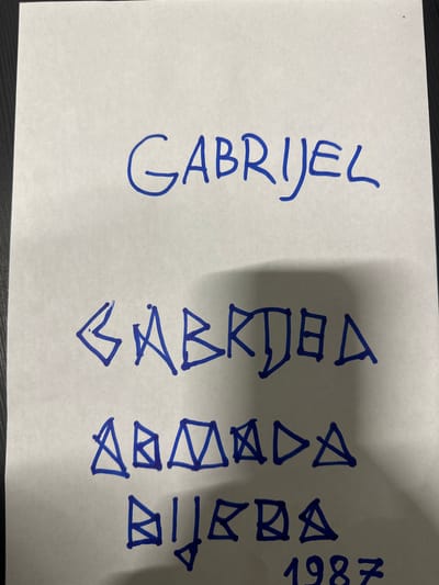 The image displays a piece of white paper with blue handwriting. At the top, the name "GABRIJEL" is written in block capital letters. Below this, in a more stylized font, is another version of what appears to be "GABRIJEL," with some letters resembling triangles or geometric shapes. Further down, there are two more lines of text written in the same stylized manner, followed by the year "1987" in block capitals. The background appears to be a dark surface, possibly a table or desk. The image is a close-up of the paper, and no context regarding its location in Schweitenkirchen, Germany, is provided within the visual information.