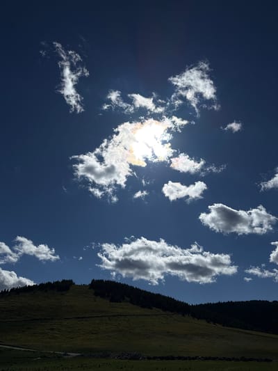 This image captures a dramatic sky filled with luminous white and grey clouds against a deep blue sky. The sun is partially obscured by a large cloud cluster, casting bright rays and colorful iridescent hues. Below the sky, a dark, tree-covered hillside slopes upward, its crest lined with dense evergreen trees. The lower part of the hill is a shadowed, grassy expanse. In the foreground, a strip of dark green grass and a hint of a dirt path suggest a rural or mountainous landscape. There is no indication of the city of Gela, Bulgaria, in this image; it appears to be a natural landscape scene.