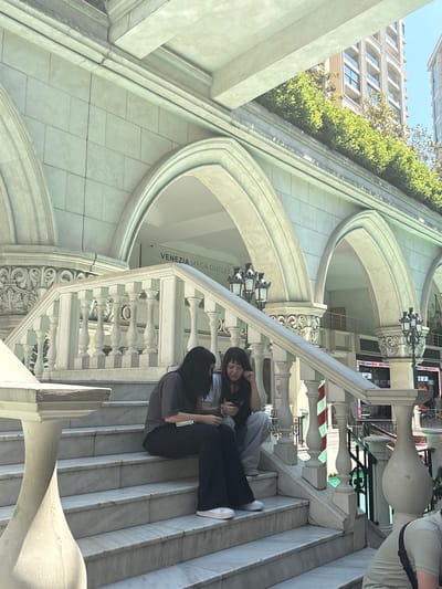 The image depicts a detailed scene at the Venezia Mega Outlet in Gaziosmanpaşa, Turkey, showcasing Venetian-inspired architecture under natural daylight. A grand staircase, constructed from light-colored marble with distinct veining, occupies the central and foreground space. The staircase features elaborate white balustrades with decorative posts and balusters.
Two women are seated on the marble steps. The woman on the left wears a dark grey short-sleeved top, dark-colored flared trousers, and white sneakers. She is looking down, engaged with an object held in her hands. To her right, another woman is seated, wearing a light-colored t-shirt, light blue jeans, and white sneakers. She is actively using a mobile phone, holding it in both hands while resting her chin on her left hand. A third individual, partially visible at the bottom right, wears a light tan t-shirt and has a dark strap across their shoulder.
The background features an expansive building facade characterized by multiple large, rounded arches and decorative columns, constructed from light-colored, block-patterned material. Ornate capitals adorn the columns, and two black, multi-lamp streetlights are mounted on the arch supports. Above the arches, a strip of vibrant green hedges suggests a rooftop garden. Beyond this greenery, modern, multi-story buildings with numerous windows are visible against a clear blue sky. A white sign with black lettering, partially visible behind the arches, reads "VENEZIA MEGA OUTLET." Subtle structural details, such as a thin crack in the stonework above one of the arches, are discernible. Below the stairs and balustrade on the right, a red and white striped pole, reminiscent of a Venetian mooring post, stands next to a glimpse of a blue-green canal or waterway.