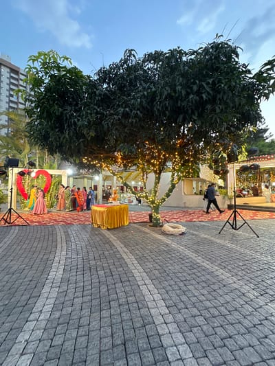 The image shows an outdoor event in Nashik, India, likely a wedding reception. The location is a paved area with gray cobblestones. A large tree is wrapped with fairy lights. In the background is a structure, also decorated with lights. A rectangular arch with a red heart-shaped decoration is visible. Some individuals are dressed in traditional Indian clothing and saris. A small gold-covered table is situated nearby. A man walks past the main structure and several sound and lighting stands are also present. The sky is partially cloudy.