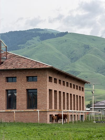 In Shabdan, Kyrgyzstan, a red brick building with a terracotta tile roof stands prominently, featuring a row of rectangular windows and a decorative brickwork facade. Two horses, one adult and one foal, are visible in front of the building, near a white fence. The environment is rural, with rolling green hills and a forest in the background under a cloudy sky. A utility pole is situated to the right of the building, and a smaller building with a dark roof is visible in the distance.