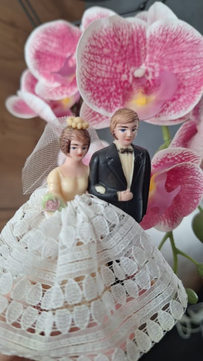 The image shows a close-up, eye-level, indoor, medium shot of a vintage plastic wedding cake topper depicting a bride and groom, placed in front of several artificial pink and white orchid flowers. The bride is on the left, wearing a yellow-toned strapless gown with a white lace overlay on her skirt. She has a veil and a small pink and green bouquet. The groom is on the right, dressed in a black tuxedo with a white shirt and a black bow tie. Both figures have blue eyes and light brown hair. The background is blurred, with the orchids being the most prominent feature. The overall lighting is soft, highlighting the details of the cake topper and flowers. The image does not provide any information about Schwaikheim, Germany, or any specific location within the city.