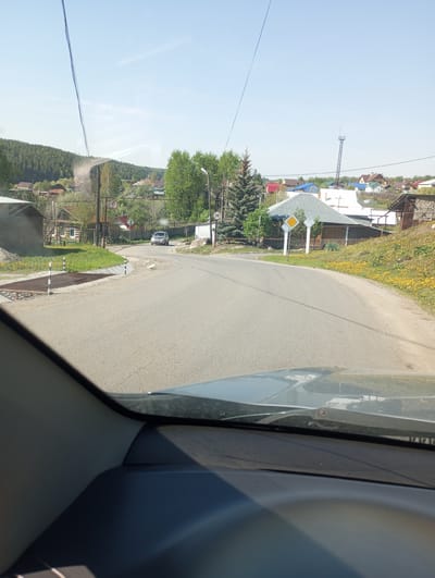 The image, taken from the interior of a vehicle, presents a view of a road in the city of Кодинка, Russia. The road curves slightly to the right, and the asphalt surface appears well-maintained. In the distance, a silver car is visible. To the left of the road, there is a cattle grate and a structure resembling a covered shed. On the right side of the road, there is a yellow diamond-shaped road sign. Beyond the road and adjacent to the buildings, a field of yellow flowers stretches across the landscape. The surrounding environment includes scattered houses and trees, with forested hills in the background. Overhead, power lines extend across the sky.