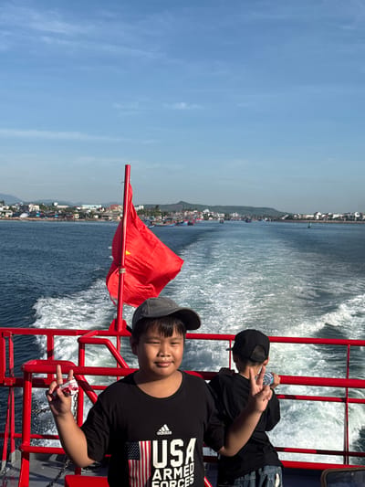 Two young boys are on the back of a boat, sailing on the sea. The boy in the foreground, on the left, is wearing a black t-shirt with an "USA Armed Forces" graphic and a grey baseball cap. He is making a peace sign with his left hand, which is holding a small item wrapped in white paper. The boy in the background, on the right, is wearing a black t-shirt and a black baseball cap. He is also making a peace sign with his right hand and holding a clear plastic water bottle. Both boys appear to be looking towards the camera.

Behind them, the boat's wake is visible in the choppy blue water. A red railing runs along the edge of the boat. A red flag on a pole is positioned behind the boy in the foreground. In the distance, a coastline with buildings and hills can be seen under a bright blue sky with some scattered clouds. This coastline is part of Tinh Khe, Vietnam. The overall scene depicts a sunny day at sea, with the boys enjoying their boat ride.
