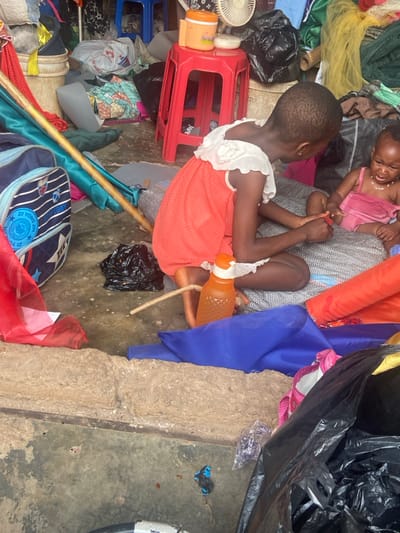 Two young children sit amidst clutter. One child in a pink outfit is held by another. The other child, wearing an orange dress with a white collar, sits on a red stool. An orange water bottle sits near her legs. Various items surround them, including clothing, a black trash bag, a blue bag, and a wooden pole. The setting appears disorganized and possibly makeshift.