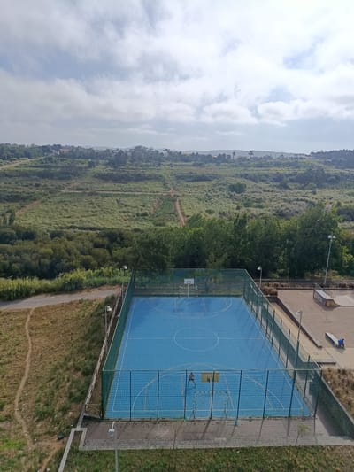 The image shows an outdoor basketball court with a blue surface and white markings, located in Algueirão-Mem Martins, Portugal. The court is enclosed by a green fence and surrounded by dry grass and vegetation. In the background, there are trees, hills, and scattered buildings, suggesting a suburban or semi-rural setting. The sky is cloudy. One person is visible on the court, appearing to be playing basketball. To the right of the basketball court, there is a skate park with ramps and a person sitting on a bench. The scene is captured from an elevated perspective, possibly from a building overlooking the area.