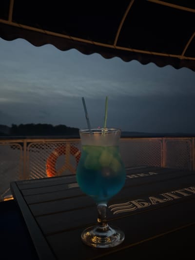 A cocktail with blue and green liquid sits on a dark wooden table. Two straws, one blue and one green, protrude from the glass. In the background, a life preserver with orange and white stripes is attached to a railing. Beyond the railing, a dark, tree-lined shore is visible under a cloudy sky. The overall lighting suggests dusk or a dimly lit evening. The table has gold lettering on it, which appears to be Cyrillic.