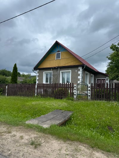The image depicts a grey, two-story house with a red roof and yellow siding under a cloudy sky. A dark brown wooden fence encloses the property, with a small, ornate metal gate in the center. The yard is covered in green grass with scattered white and yellow wildflowers. A concrete structure, possibly a ramp or platform, is visible in the foreground. A tall, slender evergreen tree stands to the left of the house, and other trees and greenery are visible in the background. The house has several windows, and what appears to be a house number "73" is visible on the exterior wall. The scene suggests a rural or suburban residential setting in Крэўскі сельскі Савет, Belarus. There are no people or animals visible in the image.
