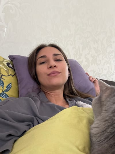 The image presents a woman reclining on a bed. She rests on a purple pillow placed on a bed with a patterned yellow and green cover. She is wearing a grey sweatshirt. A grey cat is near her head. The wall behind the bed has a light patterned wallpaper. The location is in Zanevka, Russia.