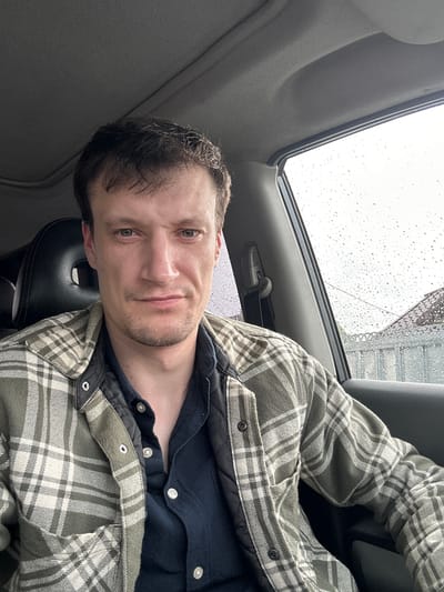 This is a selfie taken by a person inside a car. The person, a male with short brown hair and a goatee, is wearing a navy blue shirt and a green and white plaid jacket. He is looking towards the camera with a slight smile. The car interior is visible, with a grey fabric on the ceiling and a black leather seat. Through the car window, a grey, overcast sky with raindrops is visible, indicating rainy weather. The background outside the window is blurred but suggests an urban or industrial setting with some structures. The image appears to be taken during the daytime, given the brightness of the sky. There is no visible text in the image. The overall mood is casual and relaxed.