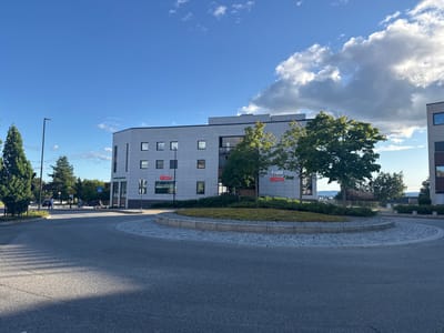 The image shows a view of a modern office building in Jessheim, Norway, with a roundabout in the foreground. The building has a light gray facade with numerous windows and the logos of "sigdal" and "aktiv" are visible on its side. A circular green area with a cobblestone border is situated in the center of the roundabout. Trees are planted around the building and on the island. In the background, a bright blue sky with scattered clouds can be seen. A few people and vehicles are visible on the street in front of the building, and a lamppost stands on the left side of the road. The scene is bathed in natural daylight, indicating it was taken during the daytime.