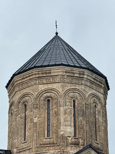 The image shows a close-up view of the domed roof and upper walls of a historic stone church, likely the Nikortsminda Cathedral in Nikortsminda, Georgia. The dome is covered in dark grey, possibly metal, shingles arranged in a faceted pattern. At the apex of the dome is a slender, multi-tiered finial topped with a cross. The stone walls beneath the dome are intricately carved with decorative bands of geometric patterns, including a prominent frieze of concentric circles, and recessed arched windows with detailed stone tracery. The stonework appears weathered, suggesting age. The sky in the background is overcast and a pale grey. The lower edge of the image shows a glimpse of a dark, angled roof structure belonging to a different part of the building.
