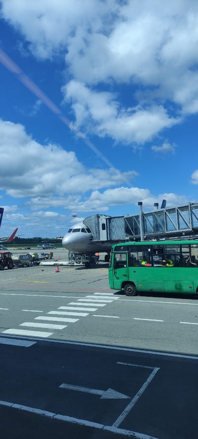The image, taken from a slightly elevated position, captures a busy airport tarmac scene under a bright, partly cloudy blue sky. The main subject is the nose section of a passenger airplane, which is connected to a gray jet bridge. The aircraft's livery is primarily white, and the words "АЭРОФЛОТ" (Aeroflot in Cyrillic) are clearly visible on its nose, along with what appears to be a part of its registration number, possibly starting with "RA-".

Parked alongside the jet bridge and aircraft is a prominent green airport bus, clearly marked with the number "10" on its side. Inside the bus, a person wearing a high-visibility yellow vest can be seen, likely ground crew or a driver. Various pieces of ground support equipment, including luggage carts, tow tractors, and other service vehicles, are scattered on the tarmac around the aircraft, indicating ongoing operations such as baggage loading or unloading.

The tarmac in the foreground features distinct white markings, including a pedestrian zebra crossing and a large directional arrow. The sky is a vibrant blue with scattered, fluffy white cumulus clouds, suggesting sunny weather during the daytime. A faint, diagonal line, possibly a reflection from the window through which the photo was taken, is visible across the upper left portion of the sky. The overall scene depicts typical airport activity at Khrabrovo, Russia.