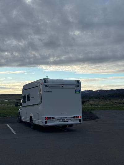 A white recreational vehicle (RV) or camper van is parked on an asphalt surface, likely a road or parking lot, with its rear facing the viewer. The RV is slightly angled, revealing parts of its side. The background features a vast, open landscape with green grassy plains extending towards distant mountains, some of which have visible patches of snow. The sky above is largely cloudy, with a mix of grey clouds and brighter areas, suggesting either late afternoon or early morning light.

There are no people visible in the image. The RV appears to be stationary, indicating a stop during travel.

Notable details include the lighting, which is soft and suggests a calm weather condition. The environment, with its expansive plains and mountainous terrain, aligns with the context of Reykjahlíð, Iceland.

Several pieces of text are visible on the vehicle:
- On the rear, "TOURINGCARS" is written horizontally, and "WEINSBERG" is written vertically.
- A European-style license plate displays "OV B21".
- The website "touringcars.com" is visible below the WEINSBERG logo.
- On the side of the RV, a sticker is present that reads "BILALEIGA AUMAN FREM Europcar", which translates from Icelandic to 'car rental'. This further confirms the Icelandic location and suggests the vehicle is a rental.