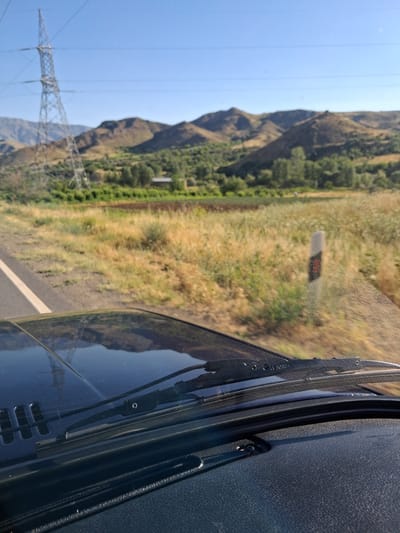The image shows a view from inside a vehicle traveling on a road, with a landscape of rolling hills and vegetation under a clear blue sky. A tall electrical pylon stands prominently on the left side of the frame, with power lines extending across the sky. The road has a white line marking its edge. The foreground is dominated by the vehicle's dashboard and windshield, with the wipers visible. The landscape in the background is characterized by dry, yellowish grass, scattered green trees, and brown, undulating hills. A small building is partially visible amongst the trees in the mid-ground. The overall impression is of a rural, somewhat arid environment. The image is consistent with a view from the road leading to the village of Getap in Armenia.