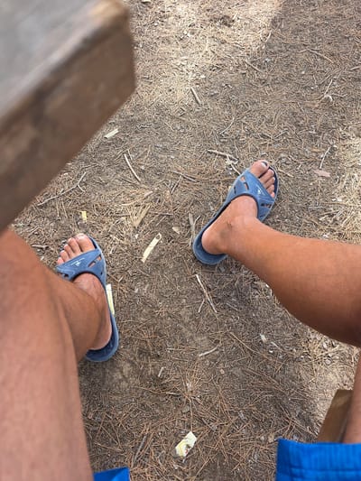 The image shows a close-up, top-down view of two legs wearing blue flip-flops, resting on a ground covered with pine needles and dry leaves, suggesting an outdoor environment like a park or forest trail. The legs are tanned, and one of the feet has painted toenails. A wooden surface, possibly a bench or table, is visible in the upper left corner. The scene appears to be set in Guardamar del Segura, Spain, a coastal town known for its pine forests and beaches.