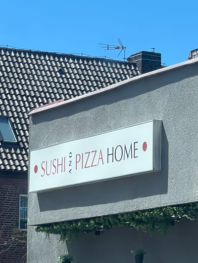 The media file shows an outdoor scene featuring a building facade and a clear blue sky. A white rectangular sign is mounted on the textured gray wall of the building. The sign displays text in a dark red font. The text reads "SUSHI PIZZA HOME" with a red dot above the "I" in SUSHI and a small "and" symbol between "SUSHI" and "PIZZA". To the left of the sign, a portion of a roof with dark, textured tiles is visible. Further left, a chimney and what appears to be an antenna are present against the bright sky. On the right side of the sign, some green foliage is growing vertically along the wall, casting shadows on the building. The lighting suggests it is daytime. The scene is static, with no people or specific activity depicted. The setting appears to be an urban or suburban environment in Ustka, Poland.