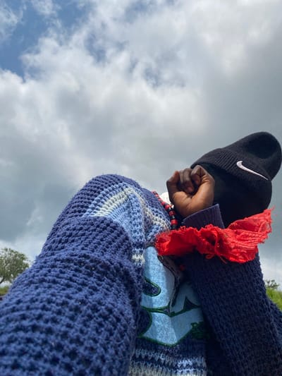 The image depicts a low-angle shot of a person wearing a dark beanie with a Nike logo, a blue and white striped knitted sweater, and a red ruffled fabric accessory around their wrist. The person's fist is clenched near their face. Red and black beaded necklaces are visible around their neck. A green graphic design with blue accents is present on the sweater. In the background, there are trees, green grass, and a cloudy sky. The provided context states the location is Ufu Esioni, Nigeria.