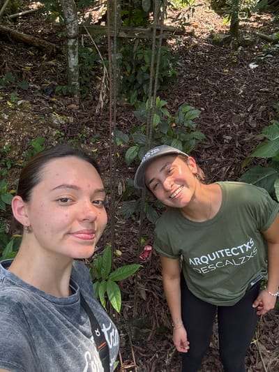 Two young women are lying on the ground in an outdoor setting, likely in Pinahuista, Mexico. The woman closer to the camera is wearing a grey t-shirt with a black strap across her shoulder. The other woman is wearing a green t-shirt with white text and a grey cap. They are surrounded by foliage, including leaves and ground cover, consistent with a wooded area. A wooden fence is also visible in the background. Both women are facing the camera and smiling.