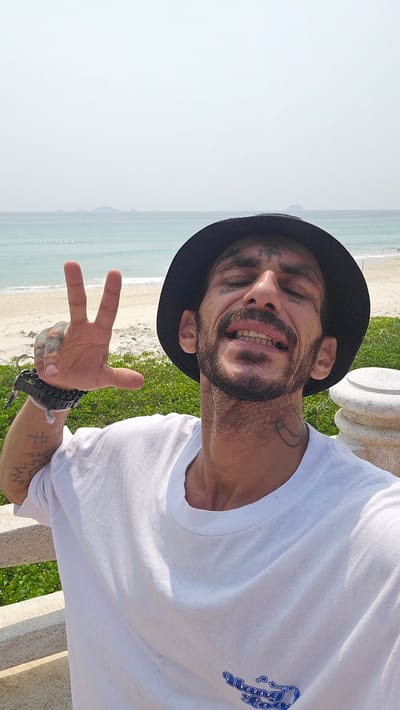 A man wearing a black bucket hat and a white t-shirt is posing for a photo with a peace sign at Bai Dai Beach in Bac Cam Ranh, Vietnam. He is sitting on a stone bench and is smiling at the camera. His shirt has a small blue graphic on the front. He has tattoos on his face, neck, and arm. In the background, there is a beach with green vegetation near the bench and a calm ocean. The sky is bright and clear.