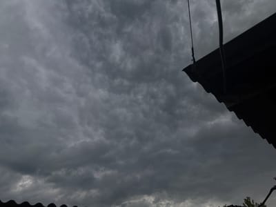 The image shows a stormy, overcast sky filled with dark, textured clouds. A portion of a dark, corrugated roofline is visible in the upper right corner, extending downwards and to the left, creating a strong diagonal line across the frame. A thin, dark rope hangs from a point above the roof, also extending downwards into the frame. In the lower portion of the image, another dark, wavy roofline can be seen, and a small glimpse of green foliage suggests trees or bushes beneath it. The overall scene evokes a sense of impending rain or a storm, with the dark clouds and muted lighting contributing to a somber or dramatic mood. The location is identified as Buky, Ukraine. No people are visible, and no text is present in the image. The time of day appears to be late afternoon or early evening, given the dark sky, but it is difficult to determine precisely due to the heavy cloud cover.